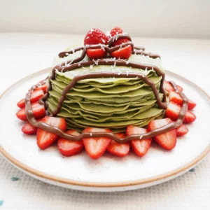 A stack of delicate, light green matcha crepes generously topped with vibrant red sliced strawberries, glossy dark chocolate drizzle, and white shredded coconut flakes, served on a speckled ceramic plate. This image beautifully illustrates a delicious matcha crepes recipe.
