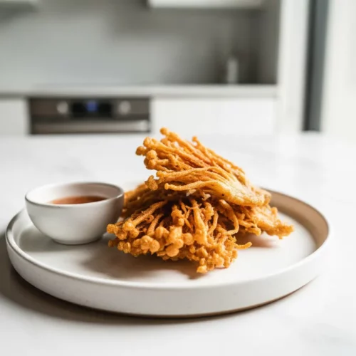 A plate of golden-brown, deep-fried enoki mushrooms piled high, showcasing their crispy texture. A small ceramic bowl of reddish-orange chili sauce sits beside the mushrooms on a white marble table.