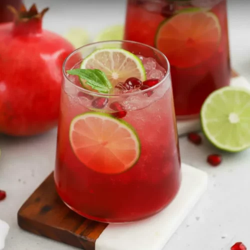 A vibrant red Holiday Pomegranate-Ginger Fizz mocktail served in a stemmed glass with crushed ice, pomegranate seeds, lime slices, and a mint sprig. The drink rests on a two-toned coaster with a whole pomegranate and additional garnishes in the background.