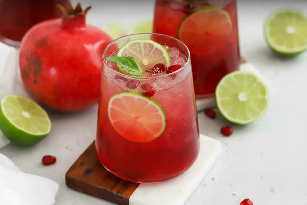 A vibrant red Holiday Pomegranate-Ginger Fizz mocktail served in a stemmed glass with crushed ice, pomegranate seeds, lime slices, and a mint sprig. The drink rests on a two-toned coaster with a whole pomegranate and additional garnishes in the background.