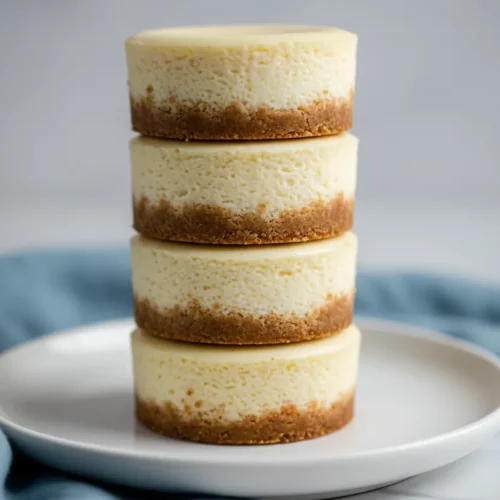 Stack of mini cheesecake bites, each with a golden-brown graham cracker crust and a creamy, pale yellow filling, arranged on a white marble surface with a blurred blue fabric in the background.