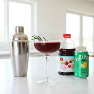 A red-colored Sleepy Girl Mocktail served in a clear stem glass, garnished with a fresh rosemary sprig. A cocktail shaker, a cherry juice bottle labeled '100% Cherry Juice', and a can of Olipop Lemon Lime soda are arranged on a mauve table cloth in the background.