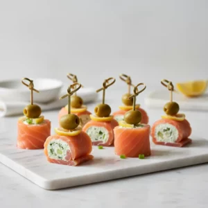 Smoked salmon and cream cheese bites, featuring thin slices of pale orange-pink salmon rolled around an off-white, herbed cream cheese filling, garnished with a green olive and a small pineapple chunk, held together by a bamboo pick. Arranged on a dark grey plate with soft, diffused lighting.