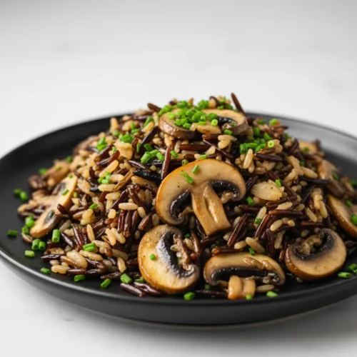 Wild rice pilaf with mushrooms served on a black plate. The pilaf features a mix of wild rice grains and sauteed mushrooms, garnished with fresh chives. The image highlights the textures and colors of the dish, including the browned mushrooms and vibrant green chives.