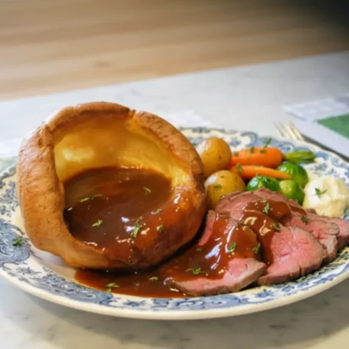 Yorkshire pudding filled with sliced roast beef and gravy, served on a decorative plate. The Yorkshire pudding is tall and golden-brown, and the beef is medium-rare.
