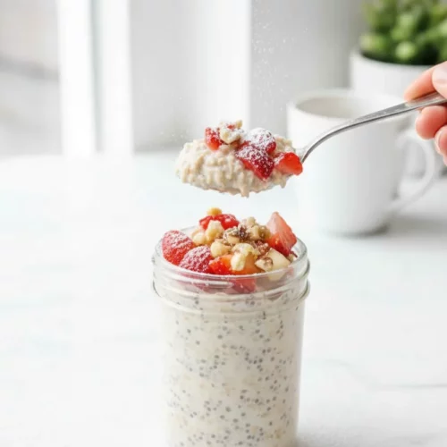 Variety of overnight oats in glass jars, each topped with different ingredients: blueberries with lemon zest, bananas and chocolate chips, coconut and chocolate chips, diced apples and walnuts, and sliced strawberries. The oats have a creamy, textured appearance.