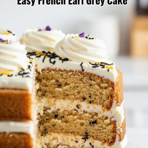 A three-layer naked French Earl Grey Cake decorated with creamy white frosting, tea leaves, and small yellow and purple flower petals, sitting on a clear glass cake stand with a light blue linen cloth beneath. The background shows a rustic wooden surface and blurred glimpses of kitchenware.