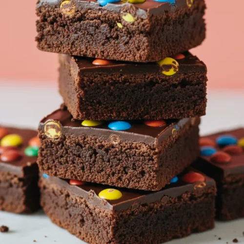 A stack of homemade cosmic brownies cut into squares, featuring a porous chocolate base, glossy ganache topping, and multi-colored candy-coated chocolates scattered across the top.