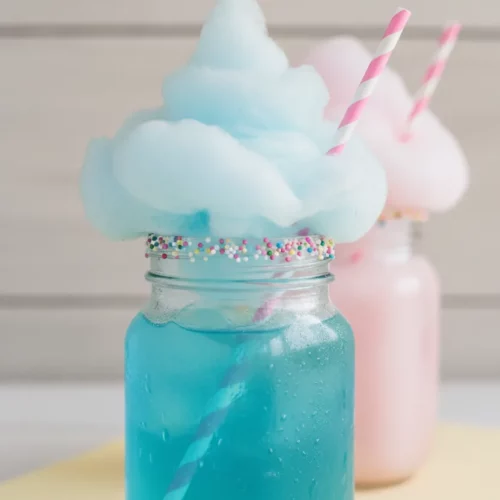 Mason jar filled with vibrant blue cotton candy soda mocktail, topped with pastel blue cotton candy and colorful sprinkles, featuring a pink and white striped straw.