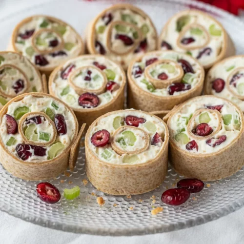 Cranberry cream cheese pinwheels arranged on a clear textured glass platter. The pinwheels are made with a light brown wrap, a cream cheese filling, red cranberries, and green celery.