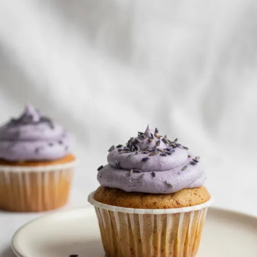 Two Earl Grey cupcakes with lavender frosting. The cupcake in the foreground sits on a white paper liner and plate, topped with a swirl of lavender-colored frosting and sprinkled with dried lavender.
