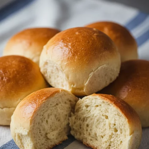 A cluster of freshly baked, golden-brown homemade dinner rolls, yeast-free, resting on a blue and white striped cloth. One roll is halved, revealing a soft, porous interior.
