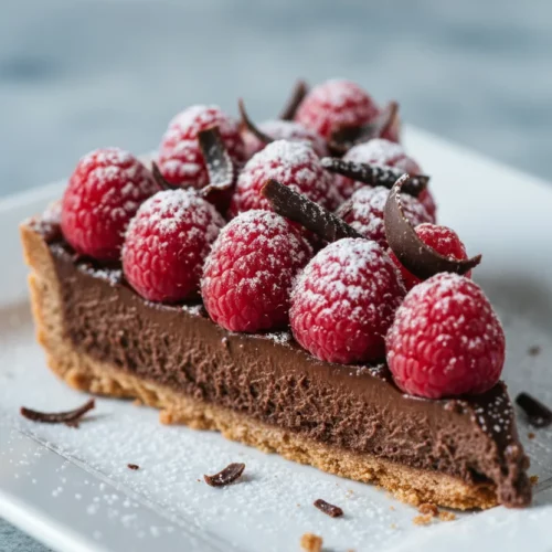 A slice of chocolate raspberry tart featuring a smooth chocolate filling, fresh raspberries dusted with powdered sugar, and dark chocolate shavings. The crust appears crumbly, served on a white plate with a fork. A dessert photography showcasing textures and bright, airy lighting.