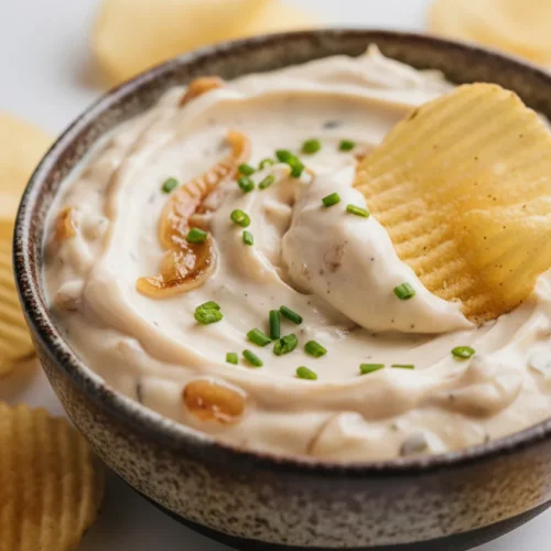 A bowl of homemade French onion dip garnished with chives, with a wavy potato chip being dipped into it. The dip is thick, creamy, and shows visible caramelized onions. Several potato chips are scattered around the bowl.