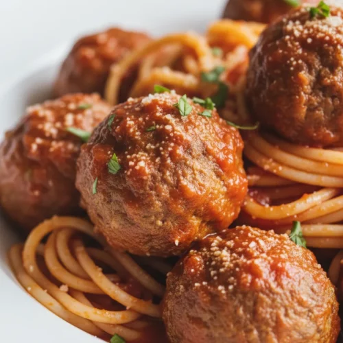 A vibrant and appetizing plate of spaghetti and meatballs, generously coated in a rich, red tomato sauce and sprinkled with grated Parmesan cheese. The meatballs have a slightly rough texture and are well-done, served in a white ceramic bowl.