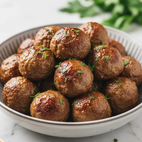 A bowl piled high with glistening turkey meatballs, coated in a semi-transparent glaze and sprinkled with fresh parsley. The meatballs are served in a grayish-white ceramic bowl on a polished marble surface, with wooden skewers in the lower left corner. This is a delicious Turkey Meatballs (Game Day Snack).