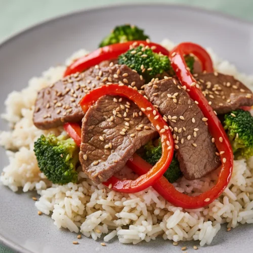 Beef Stir Fry with Vegetables served on a light grey plate. The dish features medium-well beef slices, vibrant red bell peppers, bright green broccoli, and toasted sesame seeds, all served over steamed rice, and garnished with a vintage-style fork.