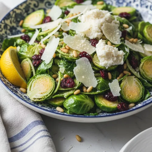 A bowl of Brussels sprouts salad sits on a white marble surface, featuring shredded sprouts, toasted pine nuts, dried cranberries, and grated parmesan cheese. A lemon wedge and a silver spoon are also in the bowl. A striped linen napkin is to the side.