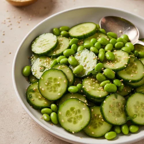 Cucumber and edamame salad in a white ceramic bowl, tossed in a light dressing with sesame seeds, presented on a beige stone surface.