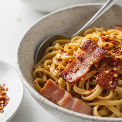 A bright and airy photo shows a bowl of fried cabbage with turkey bacon, garnished with chili flakes. The noodles have a slight sheen, and the turkey bacon pieces are irregularly cut with some caramelized edges. A steel spoon rests in the bowl, and a second bowl is blurred in the background.