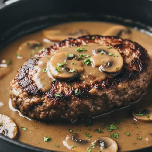 Close-up of Salisbury Steak with Mushroom Gravy in a cast iron skillet. The steak is glistening and browned, topped with chopped parsley. Sliced mushrooms are visible in the rich, brown gravy.