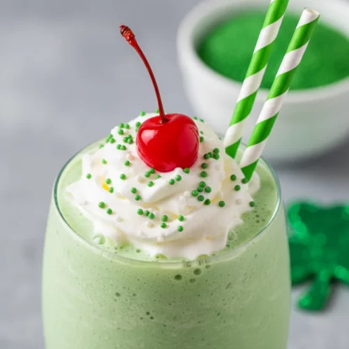 A Shamrock Shake Mint Copycat topped with whipped cream, green sprinkles, and a maraschino cherry, served in a clear glass with green and white polka-dotted straws. A green shamrock decoration and a bowl of sprinkles are in the blurred background.