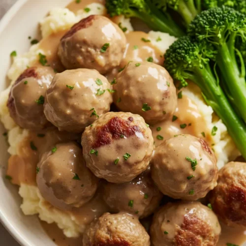A close-up, top-down shot of Swedish Meatballs (IKEA Copycat) served with mashed potatoes, gravy, and broccoli. The meatballs are coated in a light tan gravy and sprinkled with fresh parsley. The mashed potatoes are fluffy and pooling with gravy. Bright green broccoli stalks are arranged to the side.