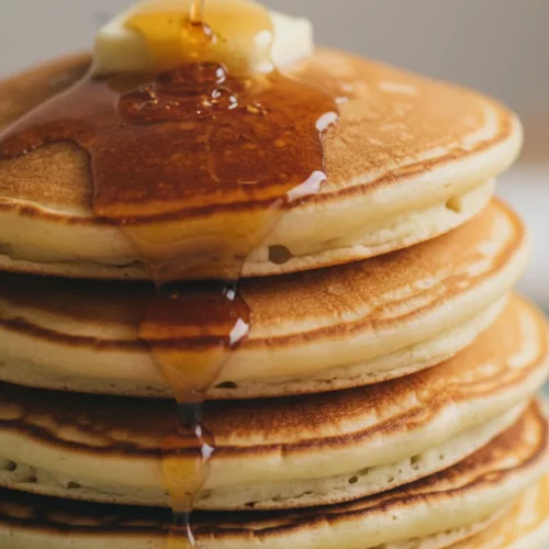A stack of golden-brown chess pie pieces with viscous maple syrup generously draped over the top. A pat of melting butter sits atop the stack, on a pale blue ceramic plate. The backdrop is a softly blurred neutral surface.