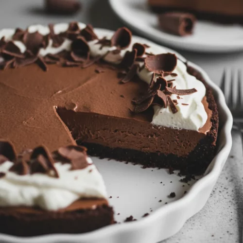 A slice of rich chocolate cream pie with a dark chocolate crumb crust, topped with soft whipped cream and dark chocolate shavings, set against a textured grey background.