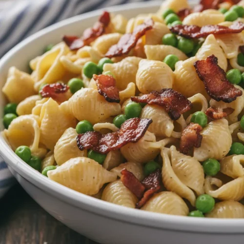 Close-up of creamy pasta salad featuring shell pasta, peas, and bacon, dressed with a creamy pasta salad dressing recipe. The ingredients are arranged in a white bowl on a rustic wooden surface with a blue and white striped linen napkin in the background.