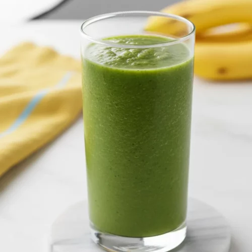 A vibrant green smoothie in a clear glass, showcasing a slightly viscous texture with visible moisture and tiny flecks of green. The smoothie sits on a round marble coaster, with a folded yellow linen napkin and blurred ripe yellow bananas in the background.