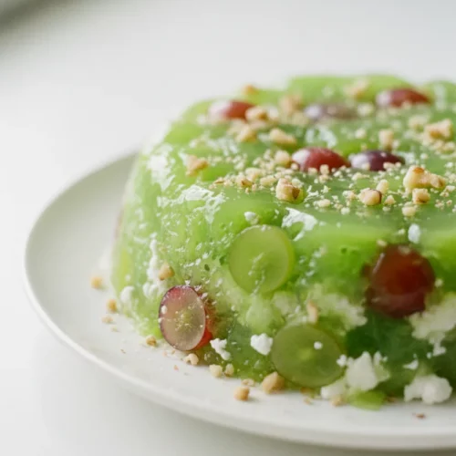 A bright, high-key food shot showcasing a lime green jello salad with a porous surface, topped with chopped nuts, translucent grape halves, scattered cottage cheese, and lightly toasted walnuts. The salad sits on a white ceramic plate on a bright white surface.