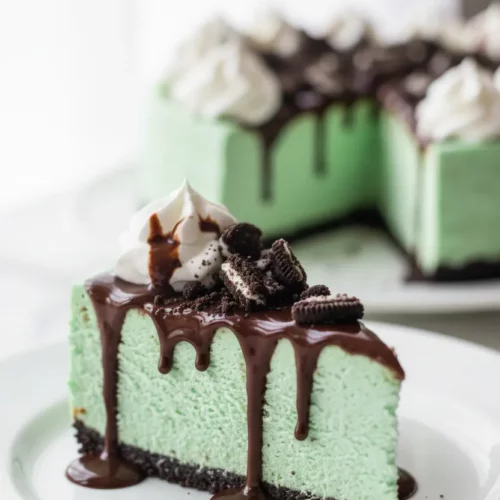 A slice of mint Oreo cheesecake with chocolate syrup dripping down the sides, topped with crumbled Oreo cookies and whipped cream. The cheesecake has a bright mint green color and sits on a white plate with a silver fork in the foreground.