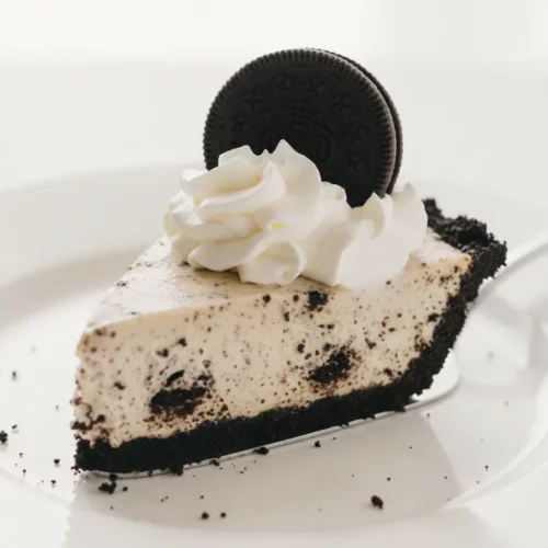 A slice of no-bake Oreo pie on a white ceramic plate, supported by a silver pie server. The pie has a creamy Oreo filling with cookie crumbs, a dark Oreo crust, and is topped with swirling whipped cream and a whole Oreo cookie.