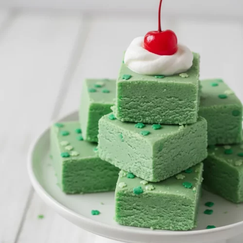 A pyramid stack of mint green shamrock fudge squares on a white cake stand, topped with whipped cream and a cherry. Green shamrock sprinkles adorn each fudge piece.