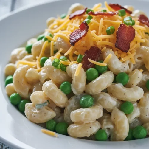 A close-up of pasta salad with peas and cheddar cheese. The macaroni shells are coated in a creamy sauce, topped with shredded cheddar, bacon bits, and chopped green onions, served on a white plate with a weathered blue wooden background.