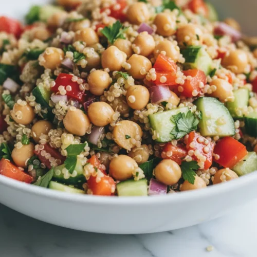 A vibrant quinoa salad with chickpeas, red bell peppers, cucumbers, and red onion in a white ceramic bowl. The salad is lightly dressed and garnished with chopped parsley, presented on a white marble surface with a tarnished silver serving spoon.