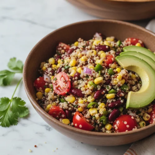 A vibrant quinoa and black bean salad served in a rustic brown bowl, featuring distinct grains of quinoa, chopped red tomatoes, yellow corn, red onion, dark kidney beans, and smooth avocado slices, with fresh cilantro and a fork. The salad is presented on a white marble surface with soft, diffused lighting.