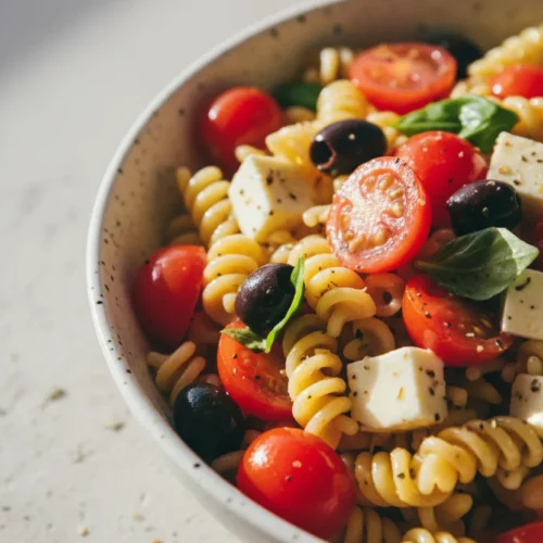 A vibrant zesty Italian pasta salad in a white speckled ceramic bowl. The salad features semi-glossy pasta, bright red cherry tomatoes, and dark purple olives, coated in a viscous dressing. A worn gold serving spoon is partially visible.