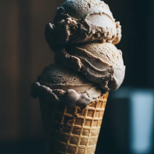 Close-up of dense, slightly grainy Blue Moon ice cream scoops in a waffle cone, dramatically lit from the right, creating strong shadows and highlights, showcasing its firm, just-scooped texture.