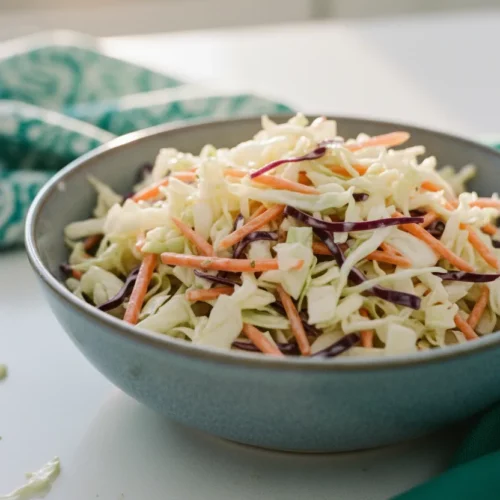 A close-up, top-down view of a vibrant no-mayo coleslaw recipe made with finely shredded white cabbage, bright orange carrot matchsticks, and deep purple cabbage, lightly coated in a creamy dressing. The salad is presented in a matte blue-grey ceramic bowl on a clean white surface with patterned and folded cloths adding subtle color.