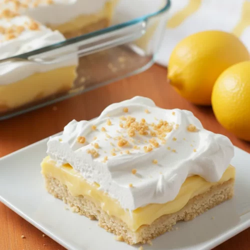 A slice of Lemon Lush Layer Dessert on a white plate, topped with whipped cream and toasted crumbs, with the rest of the dessert in a glass dish in the background. A fork is inserted into the slice. Includes whole lemons and a striped napkin.