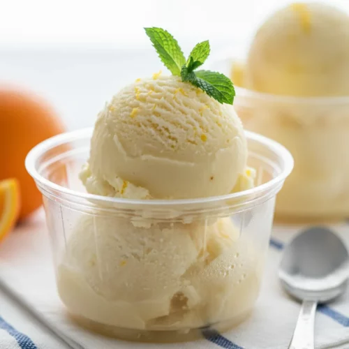 A close-up shot of scoops of pale yellow lemonade sorbet with visible zest flecks, served in a clear plastic cup and garnished with a fresh mint sprig. The sorbet has an icy texture, suggesting a refreshing and homemade quality, perfect for a hot day.