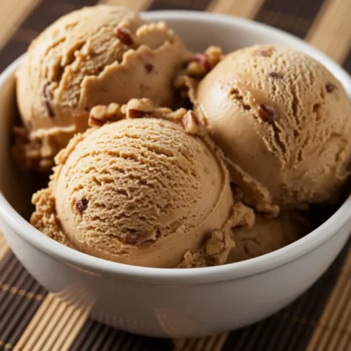 Close-up of three scoops of dense, textured maple pecan ice cream in a white ceramic bowl, with visible nut pieces, set on a striped woven placemat.