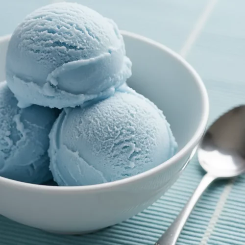 Three perfect scoops of bright yellow pineapple sorbet in a matte texture, presented in a clean white ceramic bowl. A polished silver spoon rests beside the bowl on a light blue woven placemat. The sorbet is frozen solid with defined edges.