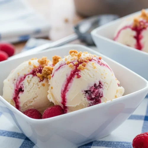 Creamy raspberry swirl cheesecake ice cream scoops in a white bowl, with visible raspberry streaks and cookie crumble. Garnished with fresh raspberries and a hint of a metal scoop.