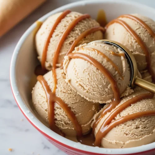 Close-up of a golden salted caramel ice cream scoop being taken with a gold ice cream scoop from a white ceramic dish with a red rim, placed on a marble surface. A waffle ice cream cone is visible in the background. The image showcases the creamy texture of the ice cream and glossy drizzle of salted caramel.