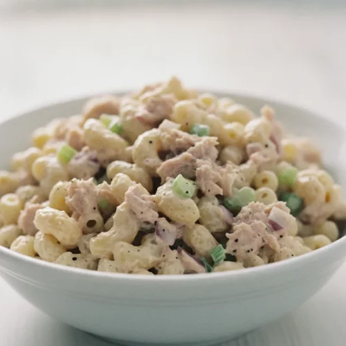 A close-up, high-key, commercial photograph of a classic tuna macaroni salad in a white ceramic bowl. The salad features creamy dressing coating perfectly cooked macaroni, mixed with flaky tuna and finely chopped celery and red onion, with visible black pepper specks. A silver fork rests beside the bowl on a light wooden surface, with a soft, blurred white and wood background.