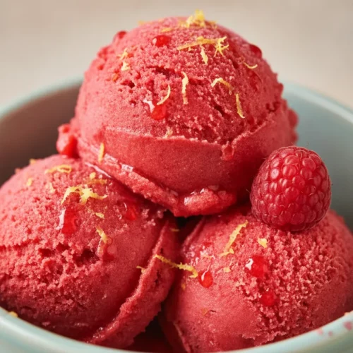 Two scoops of vibrant red raspberry sorbet in a light teal ceramic bowl, with a slightly melty, glistening, and icy texture. A single whole raspberry and fine yellow zest particles are on top. A silver spoon with a small scoop of sorbet is held above the bowl by a hand with light grey nail polish. The sorbet is presented as a refreshing 3-ingredient dessert.