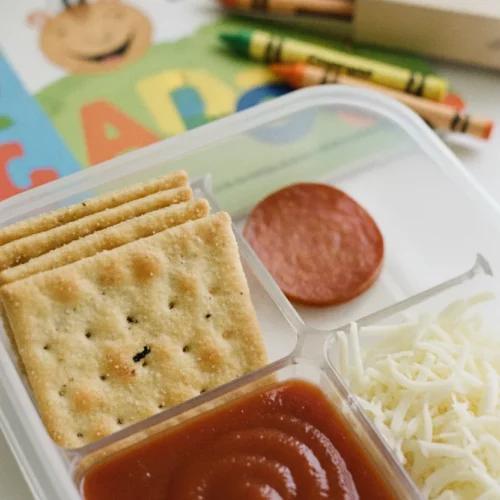 Flat lay overhead view of DIY Mini Pizza Bento, featuring golden-brown baked crackers in a divided lunchbox compartment, topped with shredded cheese, a round beef pepperoni slice, and smooth tomato sauce. Background shows colorful Crayola crayons and a children's educational book.